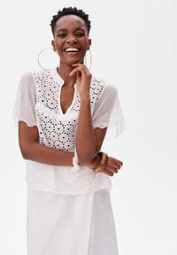 Biała bluzka z broderii anglaise z krótkimi, rozszerzanymi rękawami i falbaniastym dołem. Noszona z pasującą białą spódnicą oraz dużą drewnianą bransoletą.