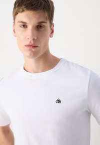 Hvid bomulds T-shirt med rund hals og et broderet sort ampersand logo på venstre brystområde, glat tekstur.