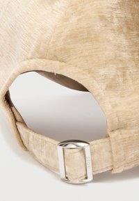 Gorra de tela beige con una superficie texturizada, que presenta una hebilla metálica plateada en la correa ajustable. La forma es redondeada con un acabado suave.