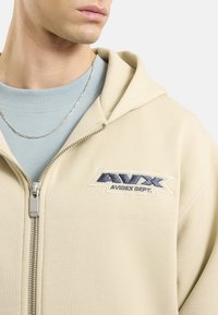 Felpa con zip beige realizzata in tessuto morbido, con una patch logo frontale, zip argentata e cappuccio attaccato. Indossata sopra una maglietta azzurra.