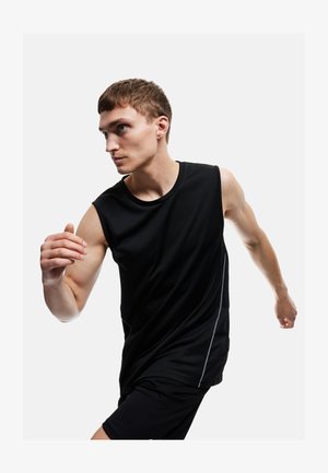 Giovane uomo con canottiera sportiva nera senza maniche e pantaloncini, posato come se stesse facendo uno sprint, guardando intensamente verso sinistra su uno sfondo semplice.