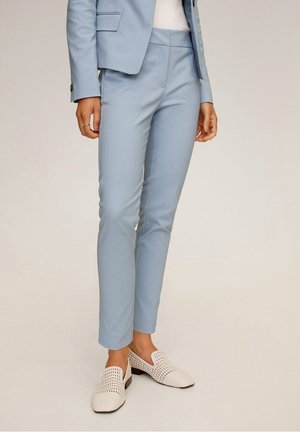 Personne portant un pantalon tailleur bleu clair, un blazer assorti, un haut en maille blanc et des mocassins tissés blancs, debout sur un fond uni.