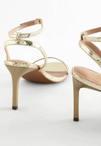 Sandali con tacco alto in metallo dorato, presentano cinturini incrociati con una finitura texturizzata, un tacco a stiletto e una soletta marrone.