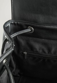 Intérieur de sac noir avec texture lisse, doté d'une poche zippée et d'œillets métalliques. Des coutures contrastées soulignent l'ouverture.