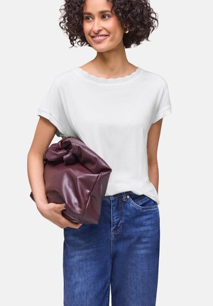 Kobieta z kręconymi włosami, ubrana w biały top z krótkim rękawem i niebieskie jeansy, trzymająca dużą złożoną bordową skórzaną torbę pod pachą.