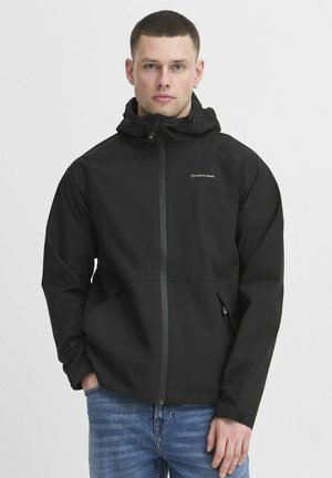 Sort vandtæt jakke med hætte, frontlynlås og logo. Har en afslappet pasform, elastiske manchetter og to sidelommer. Bæres over blå jeans.