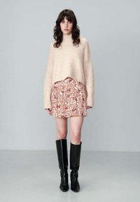 Pull en tricot beige surdimensionné avec un col montant, associé à une jupe courte à motif floral dans des tons de rose doux et d'ocre, complété par des bottes noires arrivant au genou.