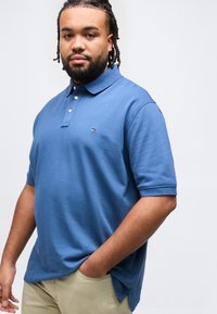 Άνδρας με πλεγμένα μαλλιά, φορώντας μπλε πόλο πουκάμισο και μπεζ παντελόνι, στέκεται με το ένα χέρι στην τσέπη, μπροστά σε απλό λευκό φόντο.