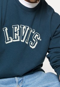 Tyrkysová mikina s velkým bílým logem "LEVI'S" v klenutém designu, vyrobená z měkkého materiálu, s žebrovaným výstřihem a manžetami.