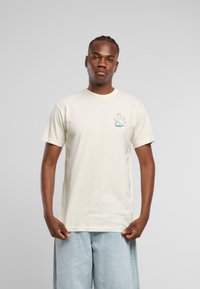 Lys beige kortermet t-skjorte laget av bomull, med et lite fargerikt grafisk motiv av en bevinget figur på brystet.