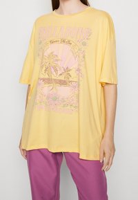 Camiseta amarilla de gran tamaño hecha de algodón, con un diseño gráfico en rosa que incluye elementos tropicales y el texto "BILLABONG Under The Sun 1973 tour."