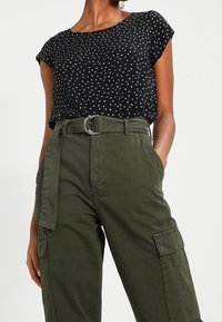 Czarna bluzka z krótkim rękawem w białe kropki, zestawiona z zielonymi spodniami typu cargo z paskiem z okrągłą, srebrną klamrą i bocznymi kieszeniami.