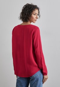 Blusa rossa a maniche lunghe realizzata in tessuto leggero, con scollo rotondo e orlo leggermente curvato. Texture liscia con vestibilità comoda.