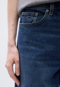 Σκούρο μπλε τζιν με διακριτική υφή. Διαθέτει μια μικρή τσέπη με λογότυπο κόκκινου, λευκού και μπλε χρώμα τος· διακοσμητικές χρυσές ραφές σε όλο το ύφασμα.