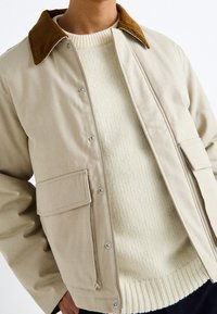 Chaqueta beige con botones a presión y un cuello de pana marrón, combinada con un suéter de punto crema con textura acanalada.