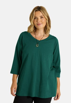 Mujer con cabello rubio lleva una camiseta verde de manga larga y un collar negro con un colgante enmarcado en oro, de pie frente a un fondo liso.