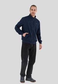 Navyblå zip-up jacka med ståkrage och elastiska manschetter, bärs med svarta jeans och grå skor. Fickor på varje sida.