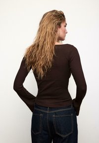 Top marrone a maniche lunghe a coste con una silhouette aderente, abbinato a denim blu scuro. Il top presenta un leggero scollo a barca e maniche svasate.