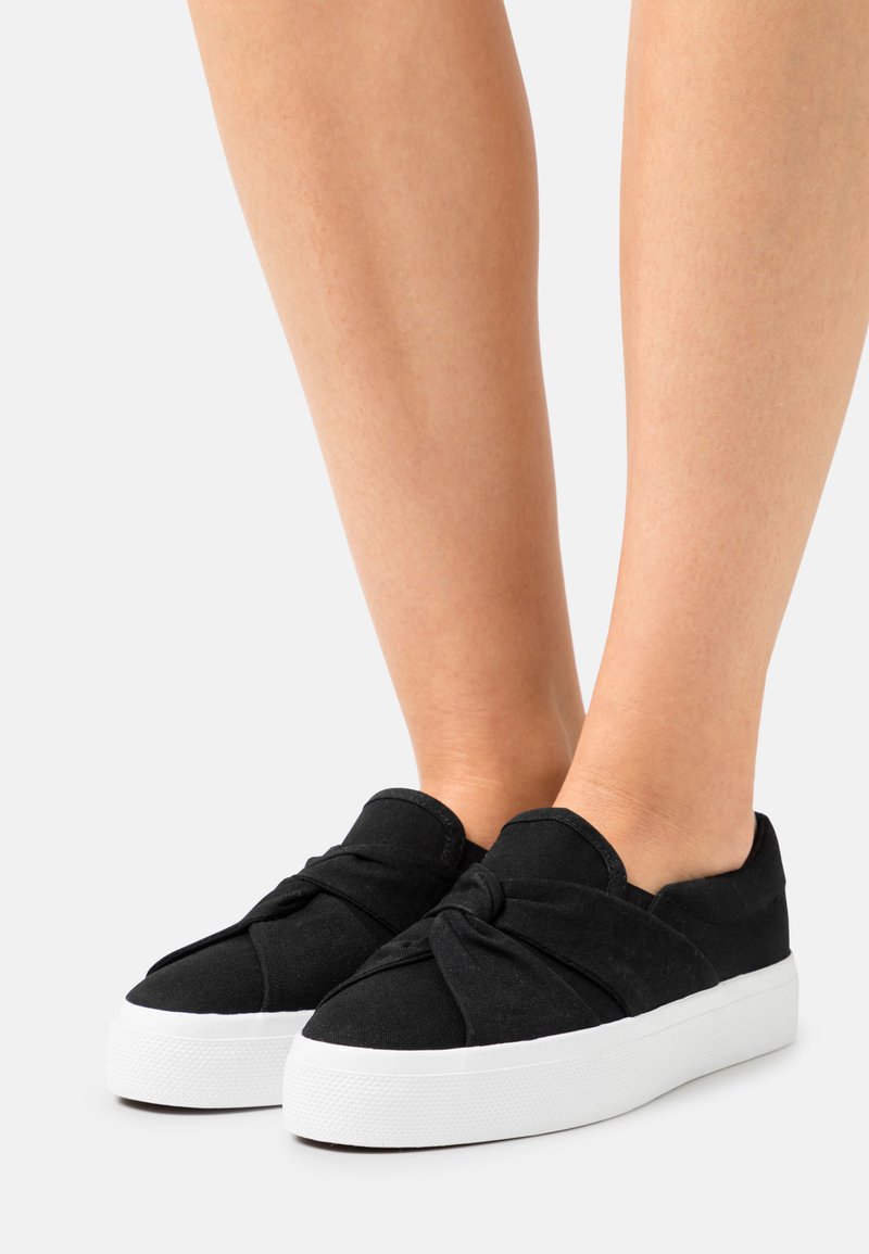 Schwarze Stoff-Slipper mit einem Knotendesign an der Vorderseite, ausgestattet mit einer weißen Gummisohle und strukturiertem Oberfläche. Minimalistischer und moderner Stil.