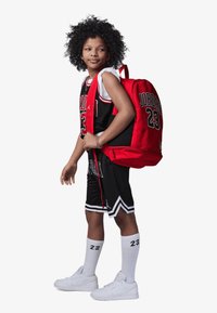 Maillot de basket noir avec des accents rouges et le numéro 23, shorts noirs assortis, chaussettes blanches avec le numéro 23, baskets blanches et un sac à dos rouge.