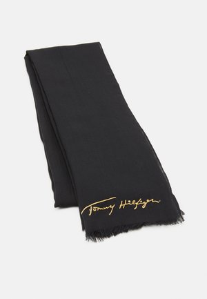 Écharpe rectangulaire noire avec bords frangés et signature brodée en or près de l'ourlet sur fond blanc.