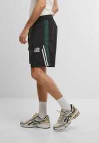 Sorte atletiske shorts med grønne sidelæg, hvide striber og et trykt logo. Matchende med sølv- og sorte løbesko.