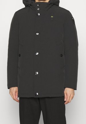 Czarna zimowa kurtka z kapturem, zapinana na guziki z przodu, z dwoma bocznymi kieszeniami i naszywką z logo na górnym lewym rękawie, noszona przez osobę.