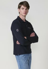 Sötétkék, hosszú ujjú pólóing bordázott textúrával, háromgombos záródással és logóval ellátott folttal a bal ujján; világoskék farmernadrággal párosítva.