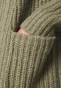 Sweter dzianinowy w oliwkowym kolorze z prążkowaną fakturą. Materiał jest gruby i miękki, posiada boczną kieszeń dla większej funkcjonalności.