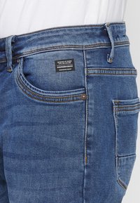 Jeans de denim azul con un diseño clásico de cinco bolsillos, costuras en contraste y una etiqueta de marca en la cinturilla trasera. Textura suave.