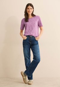 Top corto viola con motivo a foglie, abbinato a jeans blu a gamba dritta e sneaker beige, in piedi su uno sfondo neutro.