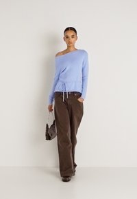 Pull bleu clair à épaules dénudées avec une ceinture nouée, associé à un pantalon ample marron. Tient un sac à main foncé avec une bandoulière chaîne.