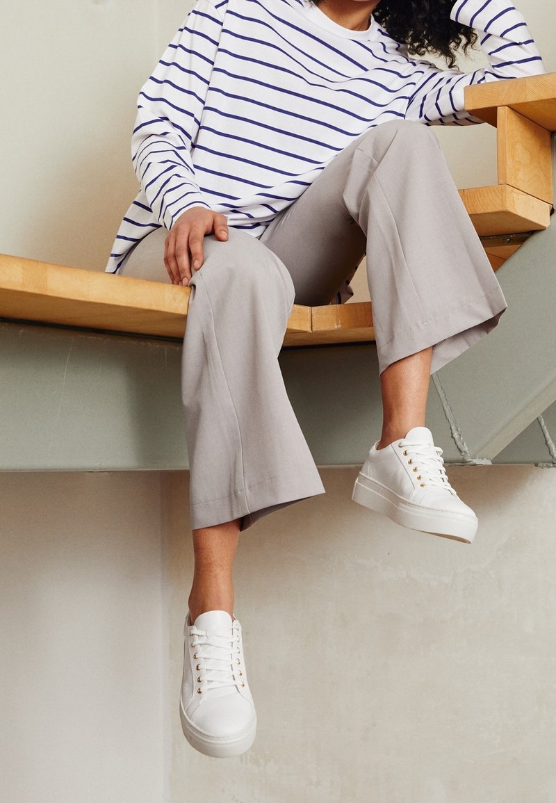 Vita sneakers med guldfärgade öljetter, kombinerade med ljusgrå vida byxor och en vit topp med blå horisontella ränder, sittande på en träbänk.