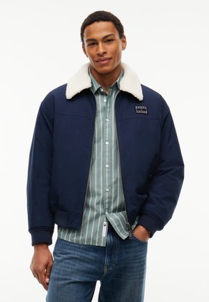 Jeune homme portant une veste bleu marine avec un col en peau lainée blanche, une chemise rayée verte boutonnée et un jean bleu, debout avec une main dans la poche.