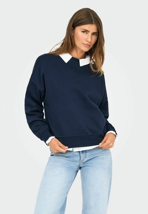 Marineblå sweatshirt med V-hals og hvid skjorte under, med lange ærmer og afslappet pasform, parret med lyseblå jeans.