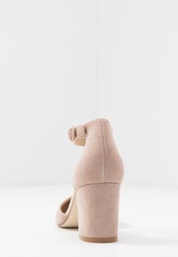 Beige semišová lodička na vysokém podpatku s blokovým podpatkem a páskem kolem kotníku, zobrazená zezadu na bílé ploše.