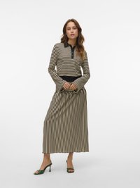 Donna con un top a maniche lunghe a righe beige e nere e una gonna a righe coordinata, in piedi con le mani unite, indossando sandali con tacco verde.