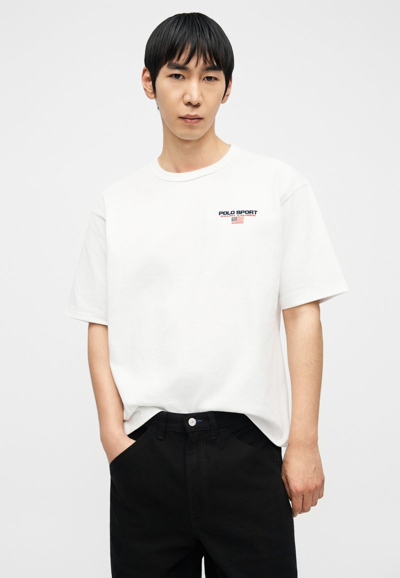 Jeune homme aux cheveux noirs portant un t-shirt Polo Sport blanc et un pantalon noir, debout les mains partiellement dans les poches, sur un fond blanc.