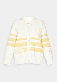 Cardigan crema con strisce gialle, caratterizzato da uno scollo a V, chiusura a bottoni e toppe con la lettera 'G', 'HWC' e il numero '43'. Texture morbida.