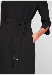 Robe noire avec une taille ceinturée, présentant une texture lisse et des manches trois-quarts. Dispose de poches latérales discrètes et de minimalismes en matière de décoration.