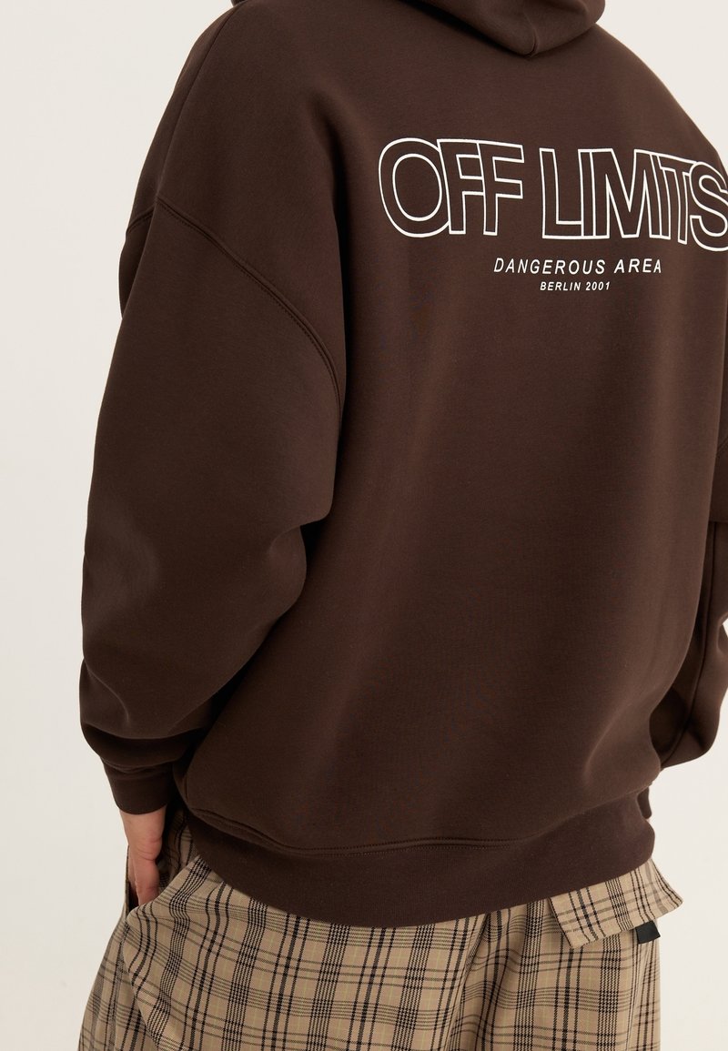 Felpa marrone con la scritta bianca "OFF LIMITS DANGEROUS AREA BERLIN 2001" sul retro; abbinata a pantaloni a quadri.
