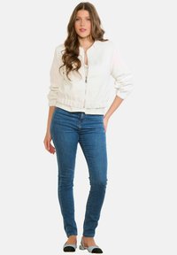 Balta zip-up bomber stila jaka ar ribotu aprocēm un apakšmalu, kopā ar zilām šaurām džinsa biksēm un melnām un gaismazilām dzīvokļu kurpēm.