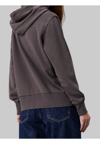 Felpa grigia con cappuccio, caratterizzata da polsini e orlo a costine. Presenta una vestibilità rilassata e una texture liscia. Indossata con jeans in denim blu scuro.