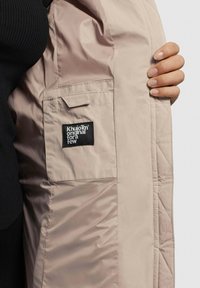 Helle beige gesteppte Jacke mit glatter Textur, ausgestattet mit einem schwarzen Logo-Patch auf der Innenseite und einer Außentasche.