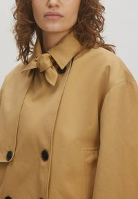 Lichtbruin trenchcoat met een gestrikt kraag, dubbele knoopsluiting en zijzakken. Gladde textuur met een klassiek ontwerp.