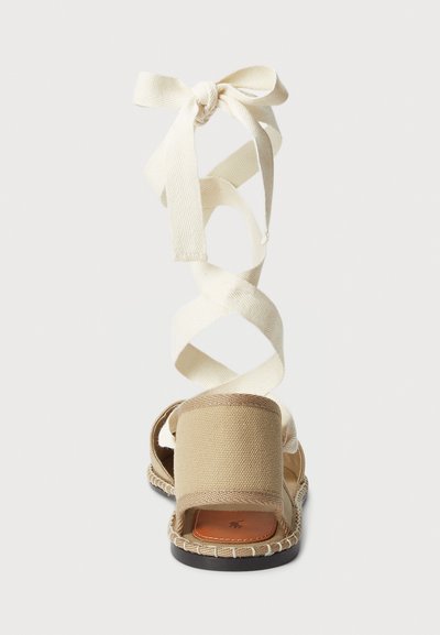 Sandalia de lienzo beige con puntera cerrada, suela plana y largas correas de algodón crema para atar alrededor del tobillo, con detalles de costura.
