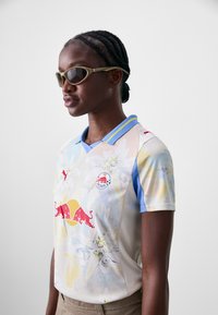 Maillot de football Puma arborant un design floral aux douces couleurs pastel, avec un col bleu et le logo Red Bull sur le devant.