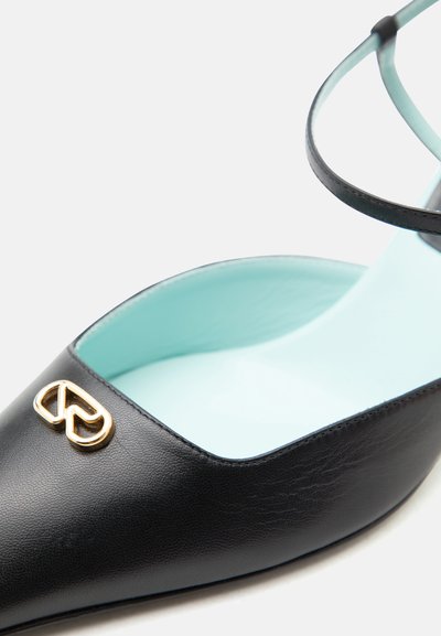 Μαύρο μυτερό επίπεδο παπούτσι με λουράκι στον αστράγαλο και χρυσή μεταλλική λεπτομέρεια σε σχήμα καρδιάς στη μύτη, με εσωτερική επένδυση σε ανοιχτό γαλάζιο χρώμα.