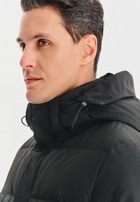 Czarna puchowa kurtka o wysokim kołnierzu z teksturowanym wykończeniem, wyposażona w kaptur i ściągacze. Zbliżenie profilu mężczyzny.