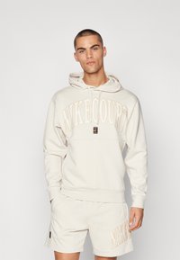Hoodie NikeCourt color beige chiaro con tasca frontale a marsupio, cappuccio con coulisse e logo ricamato, abbinato a pantaloni corti coordinati.